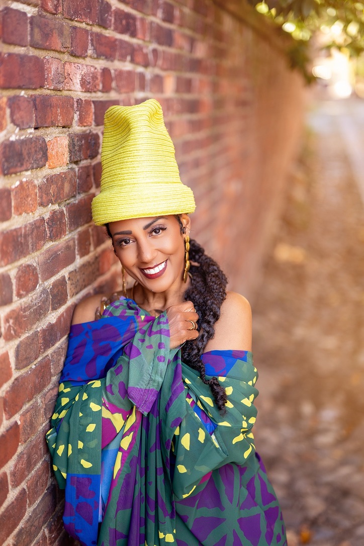 Yvette Spears in a yellow hat and a colorful wrap beside a brick wall